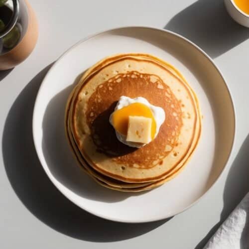 A stack of hearty pancakes topped with a dollop of cream and a pat of butter, served on a white plate. A small potted plant, a cup of syrup, and a fork with a white napkin are placed nearby—perfect for fueling up on a logging day.