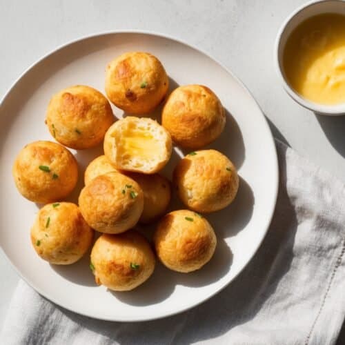 A white plate with a dozen golden-brown Pão de Queijo, one cut open to reveal a creamy filling, accompanied by a small bowl of yellow dipping sauce, on a white cloth napkin—an authentic recipe to savor.