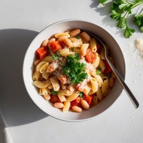 A bowl of pasta with white beans, diced tomatoes, and parsley on top, reminiscent of Pasta e Fagioli, sits next to a napkin, parsley sprigs, and a sprinkle of cheese.