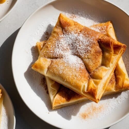 Plates of Moroccan Pastilla pastries, their golden-brown crusts dusted with powdered sugar and almonds, placed on a light-colored surface.