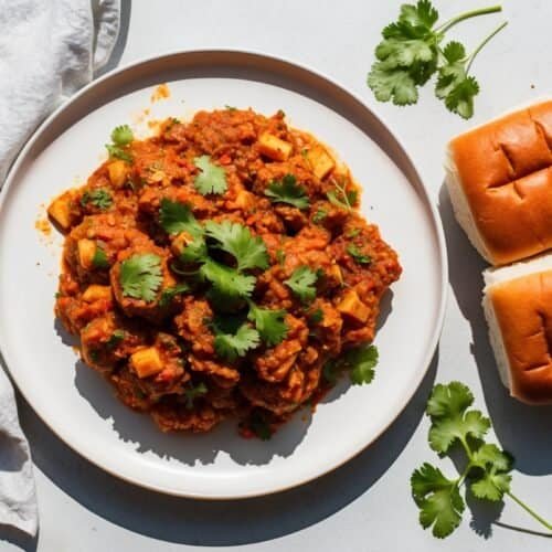 A plate of paneer curry garnished with cilantro and served with two whole wheat buns. A white cloth and more cilantro sprigs are placed beside the plate on a white surface, making this recipe a healthy twist on traditional delights.