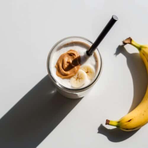 A quick, frothy smoothie with banana slices and a dollop of nut butter, accompanied by a black straw, sits next to a ripe yellow banana on a white background – the perfect nutritious protein shake.