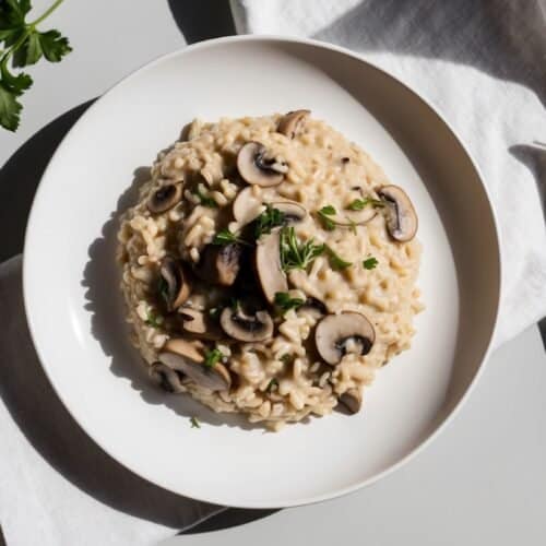 A bowl of creamy mushroom risotto garnished with parsley, placed on a white napkin with a fork beside it.