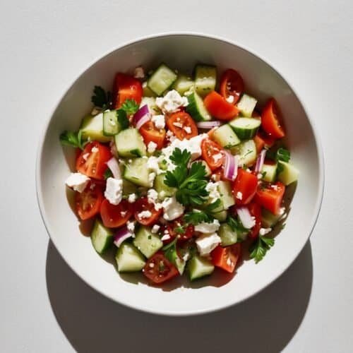 A bowl of refreshing salad containing chopped cucumbers, tomatoes, red onions, feta cheese, and parsley, placed on a white surface. This Bulgarian Shopska Salad is perfect for any occasion.