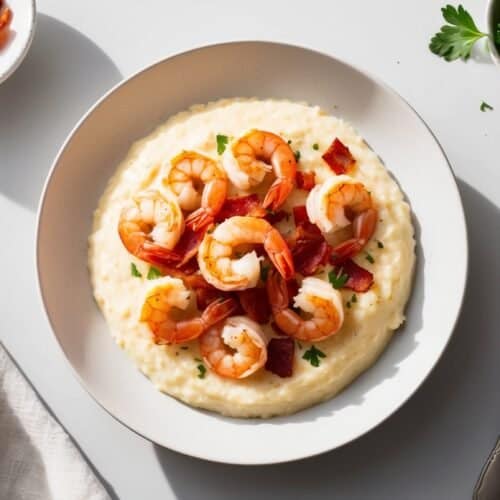 A plate of shrimp and grits topped with pieces of crispy bacon, garnished with parsley, is set on a table alongside a fork, a napkin, a small bowl of bacon, and fresh parsley. This mouthwatering recipe combines the savory flavors of the South in every bite.