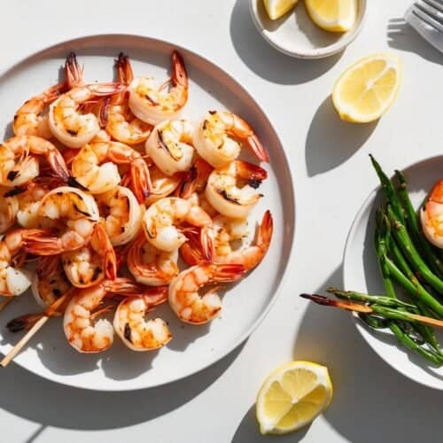 A plate of grilled shrimp with lemon wedges and a small plate of shrimp skewers and green beans, accompanied by a napkin and silverware on a white surface—an impeccable Barbie Recipe.