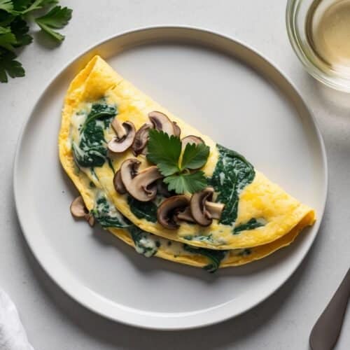 A plate with an omelette filled with spinach and topped with sliced mushrooms and parsley. A fork, a glass of white wine, and a sprig of parsley are beside the plate.