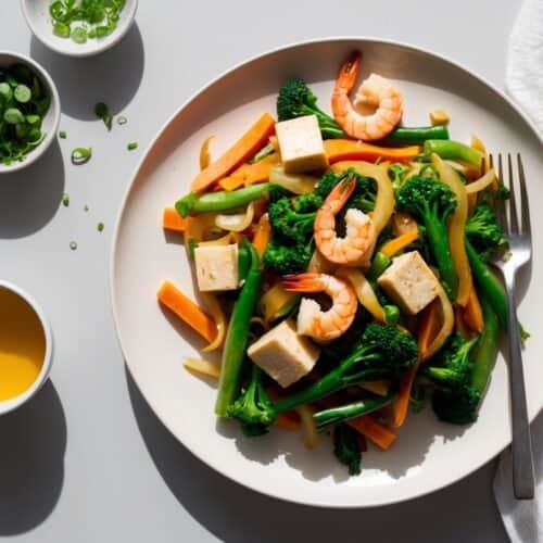 A quick and nutritious plate of mixed vegetables, tofu, and shrimp with a fork, accompanied by small bowls of chopped green onions and yellow sauce, and a white napkin on the side. Perfect for a veggie stir-fry lover!