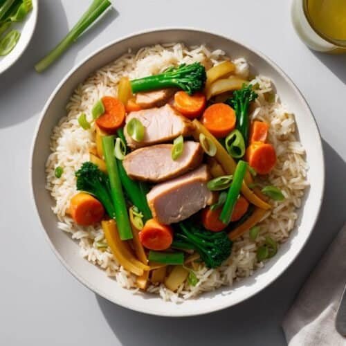 A nutritious bowl of rice topped with sliced pork stir-fry, broccoli, carrots, and green onions. A fork, napkin, glass of juice, and a small plate of green onions are beside the bowl.