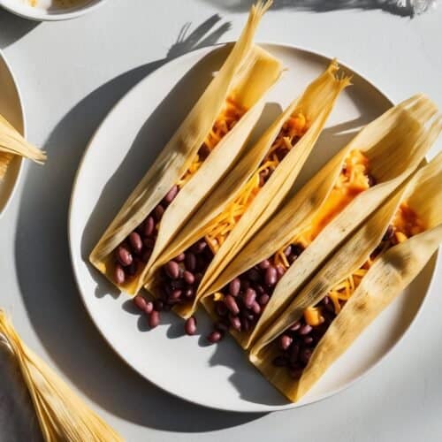 Four authentic Mexican tamales filled with beans and cheese are arranged in open corn husks on a white plate. Surrounding the plate are additional corn husks.