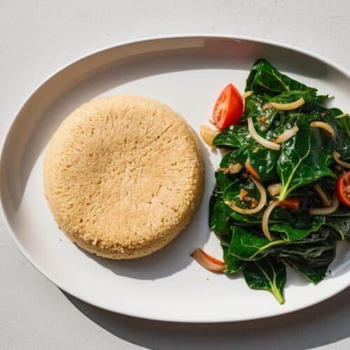 A white plate with a dome-shaped portion of fufu alongside a mixed vegetable dish featuring greens, tomatoes, and sliced onions reminiscent of Sukuma Wiki.