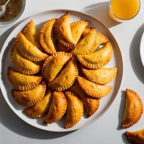 A plate of golden-brown vegan empanadas arranged in a circle on a white surface, with a glass of orange juice, a bowl of brown sugar, and a few empanadas on a smaller plate and the surface around it.