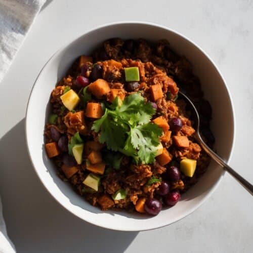 A bowl of chili topped with diced avocado, chunks of sweet potato, kidney beans, and a sprig of cilantro, with a spoon inside. This flavorful vegan dish sits on a white surface, accompanied by a cloth napkin.