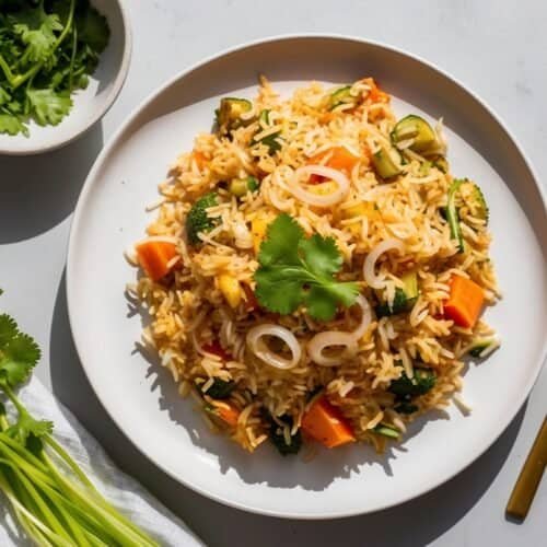 A plate of healthy vegetable biryani made with brown rice, garnished with cilantro, next to a gold fork and a small bowl of cilantro.