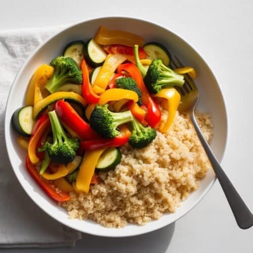 A bowl of healthy quinoa with broccoli, sliced red and yellow bell peppers, and zucchini on a white napkin, with a fork resting inside the bowl.