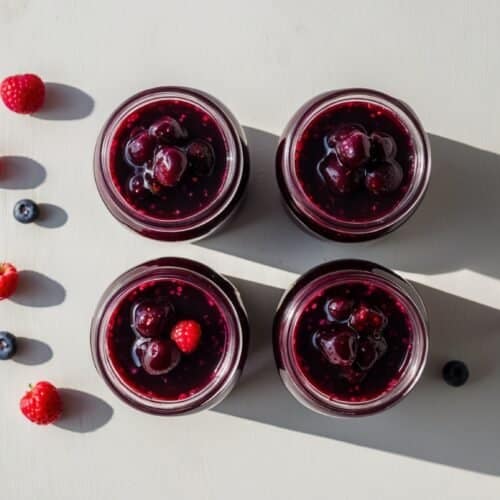 Four jars of homemade berry dessert are arranged in a square on a white surface, surrounded by scattered raspberries and blueberries. Two shadows cast to the right from the jars, adding to the charm of this wild berry delight.