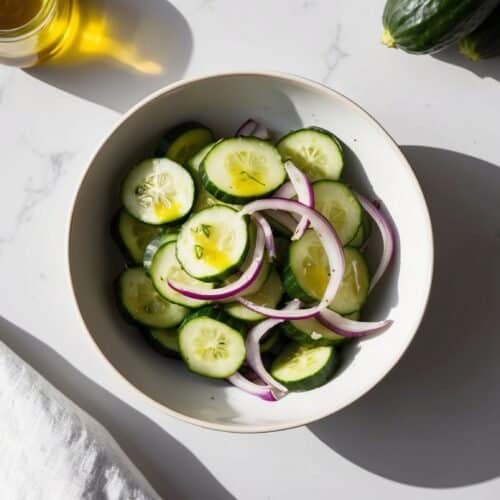 A bowl of sliced cucumbers and red onions on a white surface with a cloth, a jar of pumpkin seed oil, and two whole cucumbers nearby creates the perfect scene for a refreshing cucumber salad.