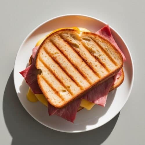 A toasted sandwich with ham and melted cheese is placed on a white plate.