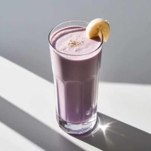 A tall glass filled with a purple, vegan smoothie, garnished with a banana slice on the rim, is placed on a surface with sunlight casting shadows—perfect for a post-workout refreshment.