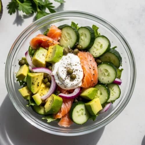 A glass bowl of cucumber salad featuring smoked salmon, avocado chunks, cucumber slices, red onion, capers, and greens, topped with a dollop of cream. An avocado half, parsley sprigs, and a small bowl of capers sit nearby.