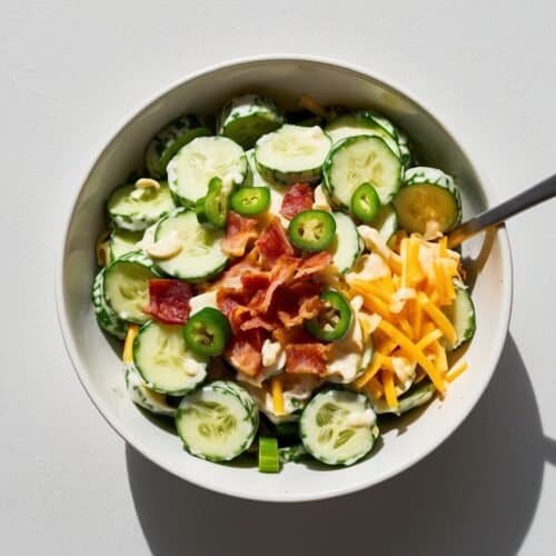 A bowl of cucumber salad containing sliced cucumber, chopped bacon, shredded cheese, jalapeño slices, and a creamy dressing reminiscent of a Jalapeño popper recipe, with a fork placed inside the bowl.