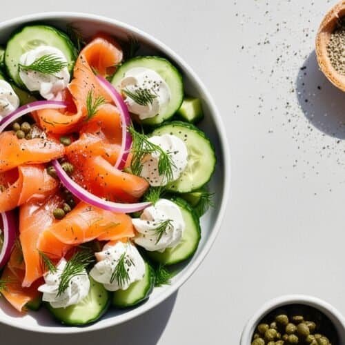 A bowl of salad with smoked salmon, sliced cucumbers, red onions, dollops of cream cheese reminiscent of a salmon cream cheese bagel, and dill garnish. Nearby are a bowl of capers, a small dish of pepper, a sprig of dill, and scattered pepper.