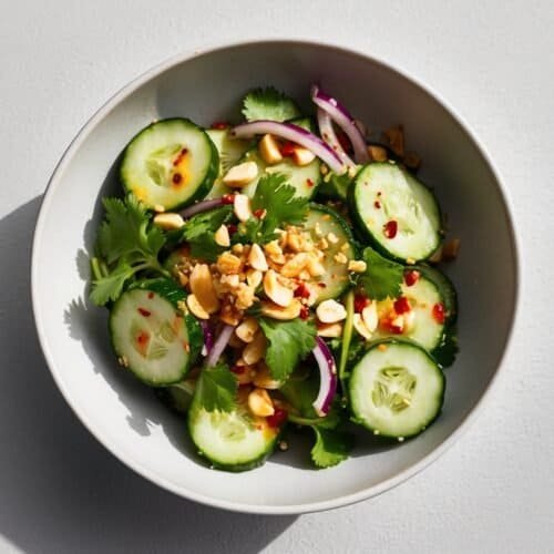 A white bowl contains a Spicy Thai cucumber salad made of cucumber slices, red onion, cilantro, and chopped peanuts, all tossed in a light dressing.