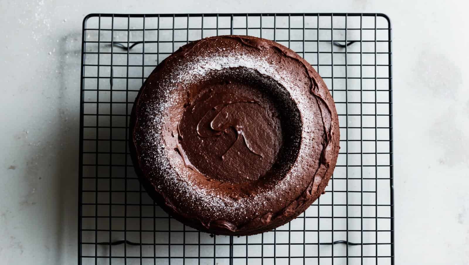 A moist chocolate cake dusted with powdered sugar rests on a cooling rack.