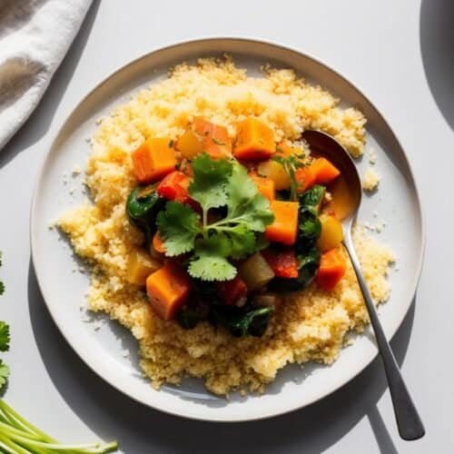 A vibrant plate of couscous topped with a colorful vegetable medley, delicately garnished with cilantro, all poised perfectly beside a spoon. A sprig of cilantro completes this delightful recipe.