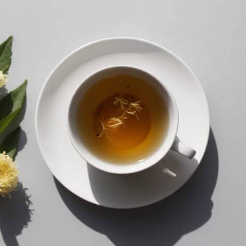 A white cup of soothing floral tea with floating flowers rests on a saucer. Beside it lies a sprig with two small yellow blooms, all elegantly set on a gray surface.