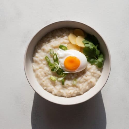 Enjoy a comforting bowl of Jok Thai rice porridge, featuring a soft boiled egg, sliced green onions, ginger, and cilantro for a fragrant finish.