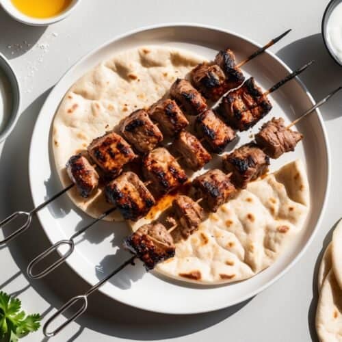 Grilled lamb kebab skewers rest on a plate with flatbread, accompanied by small bowls of yogurt sauce and parsley on a white surface, reminiscent of a street vendor's favorite recipe.