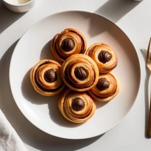 A white plate with six round pastries topped with chocolate swirls, reminiscent of a classic Chocolate Croissant Recipe, placed on a light surface next to a fork.