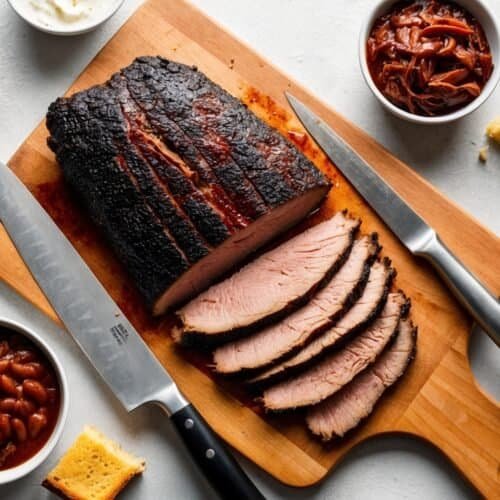 Sliced smoked brisket on a cutting board with a knife, accompanied by bowls of coleslaw, baked beans, caramelized onions, and cornbread pieces. It's a ranch recipe delight for any barbecue enthusiast.