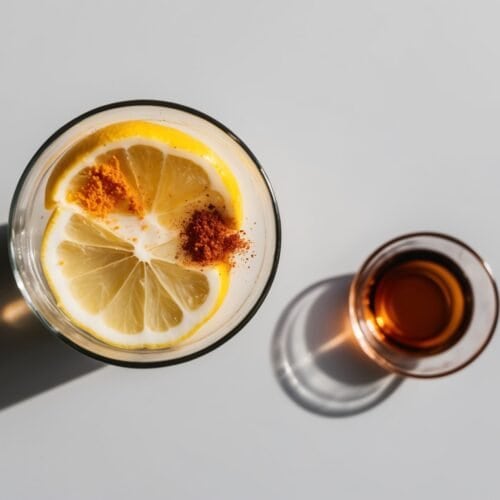 A glass of lemon water with lemon slices and spices sits next to a small glass of brown liquid on a white surface, perfect as a morning detox drink loved by many, including Beyoncé.
