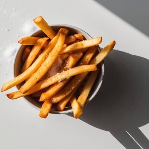 A bowl of golden, crispy French fries on a light surface, as balanced and precise as a Simone Biles routine, sprinkled with salt. Shadow cast to the right.
