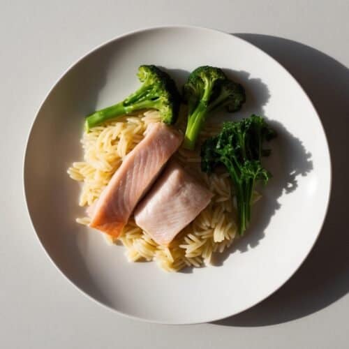 A balanced dinner featuring a plate of orzo pasta, nestled alongside two pieces of pink fish and vibrant broccoli, set against a clean white background.