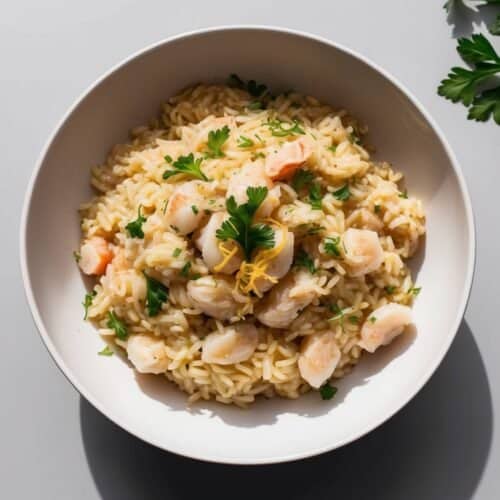 A bowl of shrimp risotto with a hint of lemon zest and a sprinkle of Parmesan, garnished with parsley on a light gray surface adorned with parsley sprigs.