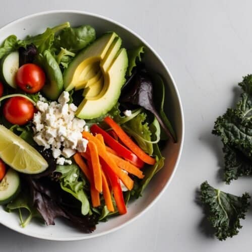 A bowl of vibrant mixed salad features avocado, cherry tomatoes, cucumber, lettuce, carrots, and feta cheese topped with a lime wedge. Beside it on a light gray surface are kale leaves ready to transform into crunchy kale chips.