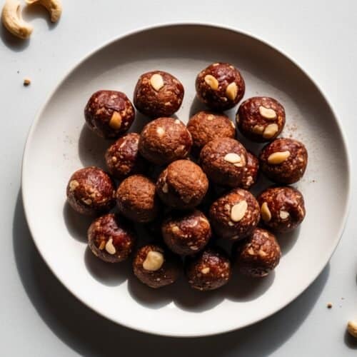 A plate of chocolate energy bombs is arranged on a white surface, surrounded by a few cashews and one date.
