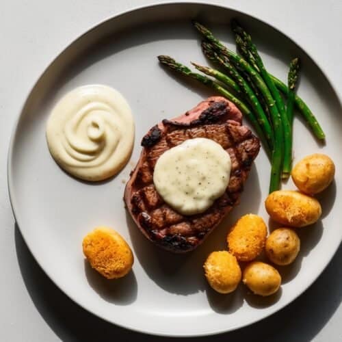 A plate with grilled steak au poivre topped with sauce, green asparagus, small Parmesan-crusted potatoes, a swirl of white sauce, and a round breaded item on a light surface.