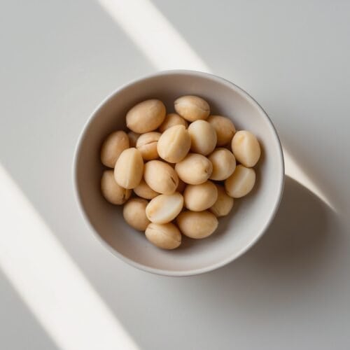 A white bowl filled with shelled peanuts, known for their brain-boosting benefits as endorsed by Andrew Huberman, sits on a light-colored surface with striped shadows cast across it.