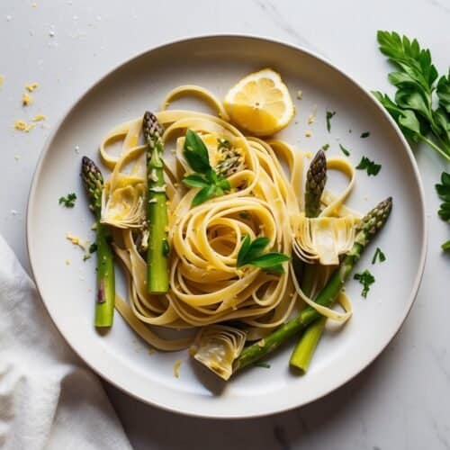 A plate of artichoke and asparagus fettuccine with lemon and herbs rests on a marble surface. A cloth napkin and parsley sprigs are artfully placed beside this delightful pasta dish.