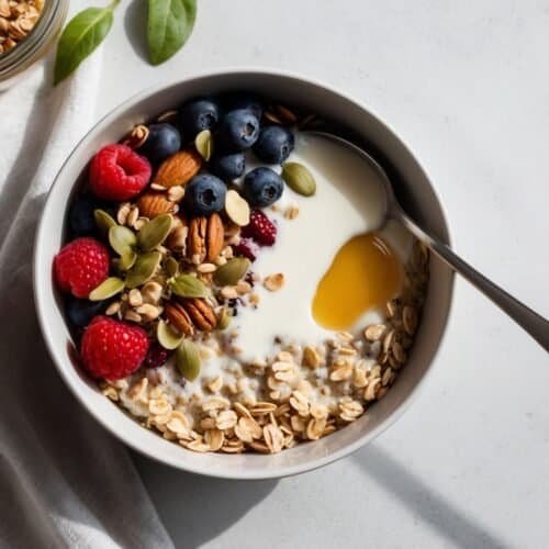 A power bowl of creamy yogurt topped with fresh raspberries, blueberries, crunchy nuts, seeds, and a drizzle of honey sits on a white surface. Beside it, a jar of granola and a cloth napkin await your next nourishing bite.