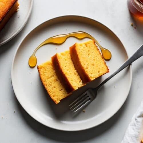 Slices of yellow cake on a plate with a drizzle of honey and a fork, reminiscent of Oprah’s Honey Cornbread, sit invitingly next to a jar of honey.