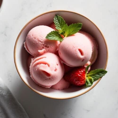 A refreshing bowl of strawberry sorbet with three luscious scoops, garnished with fresh strawberries and mint leaves, sits elegantly on a white surface.