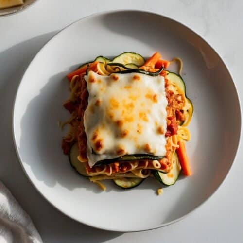 A plate of turkey lasagna with veggie noodles, topped with melted cheese, sits elegantly on a white plate. It's placed on a light surface next to a folded napkin, evoking a meal worthy of Oprah’s table.