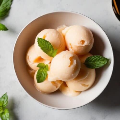 A bowl of peach sorbet infused with hints of ginger, garnished with fresh mint leaves, sits beside a spoon and a sliced peach on a light surface.