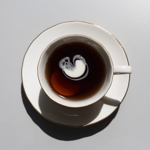 A white cup on a saucer contains a bold black coffee with a swirl of milk, viewed from above—a delightful morning ritual reminiscent of Renée’s Tea.