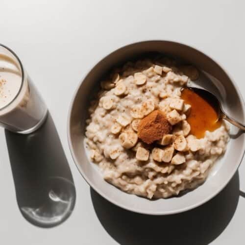 A bowl of protein-packed oatmeal topped with marshmallows and a sprinkle of brown powder sits enticingly next to a glass of milk on a white surface, perfect for a post-workout treat.